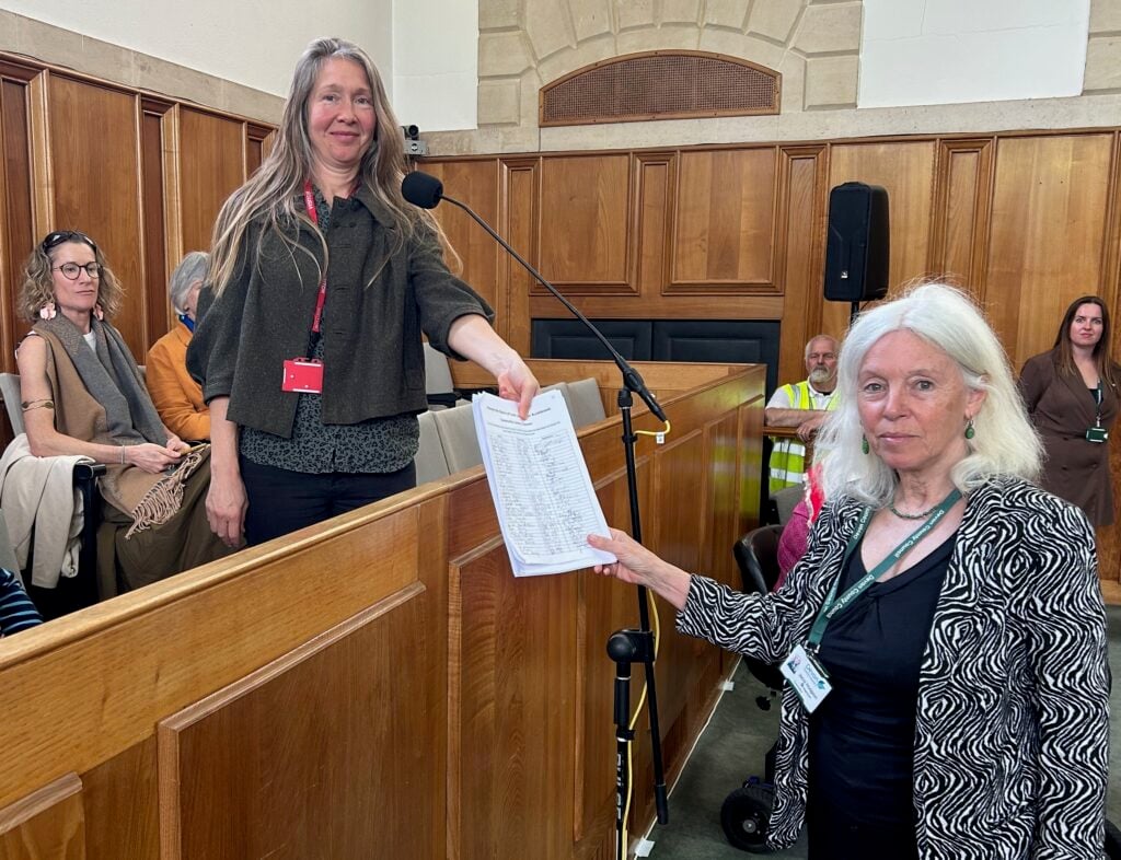 Cllr Jacqi Hodgson with Dr Sally Murrall-Smith holding a petition in the council chamber. There are other people in the public seating.