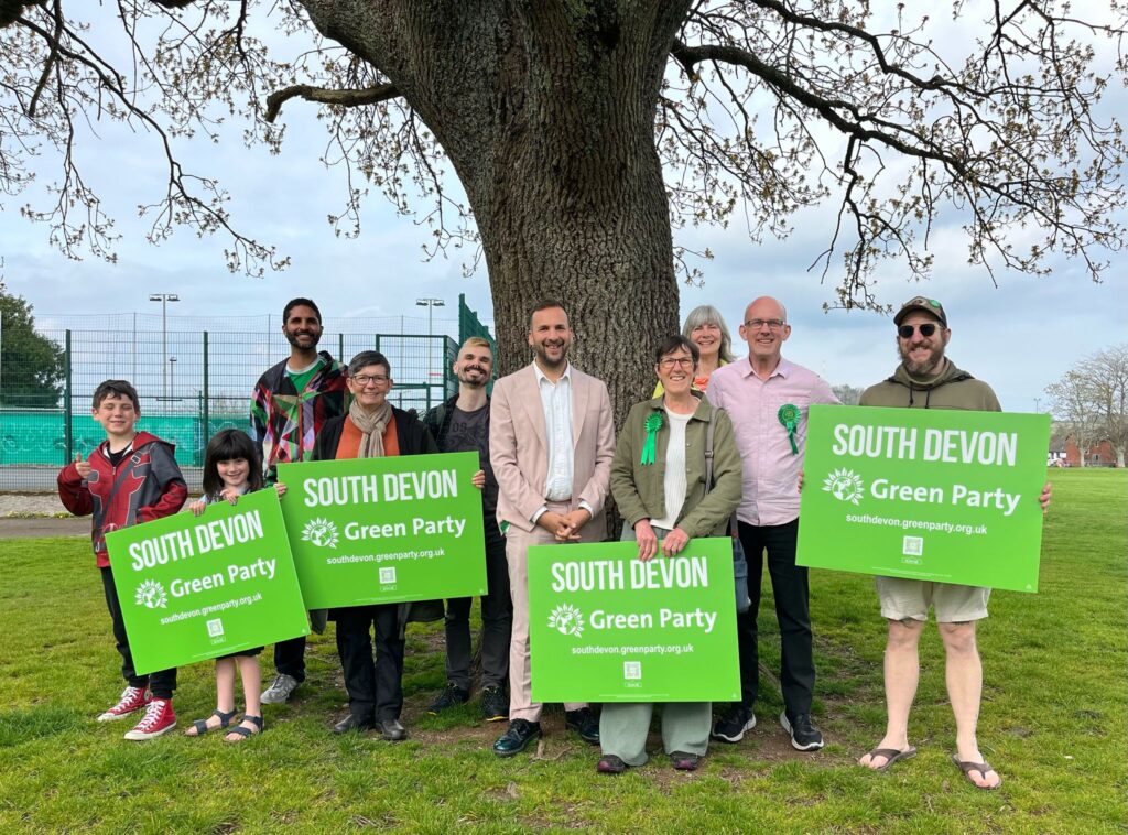 A group of people under a tree, holding south Devon Green Party banners and smiling.