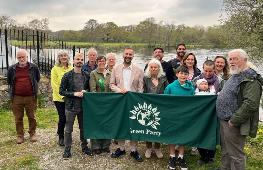 Zack Polanski and activists stand on the bank in front of the River Dart holding a large Green Party banner. There are trees on the opposite bank and a cloudy sky.