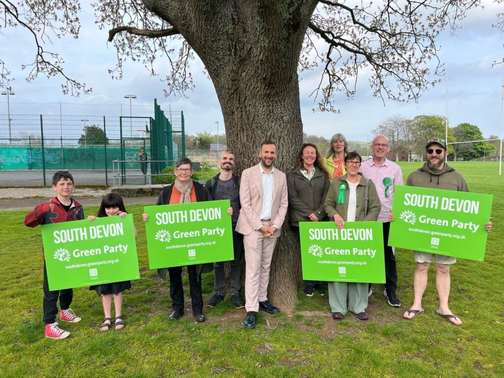 Zack Polanski stands with Green Party candidates, councillors and activists and two children under a tree in a park. They are holding South Devon Green Party banners and smiling.