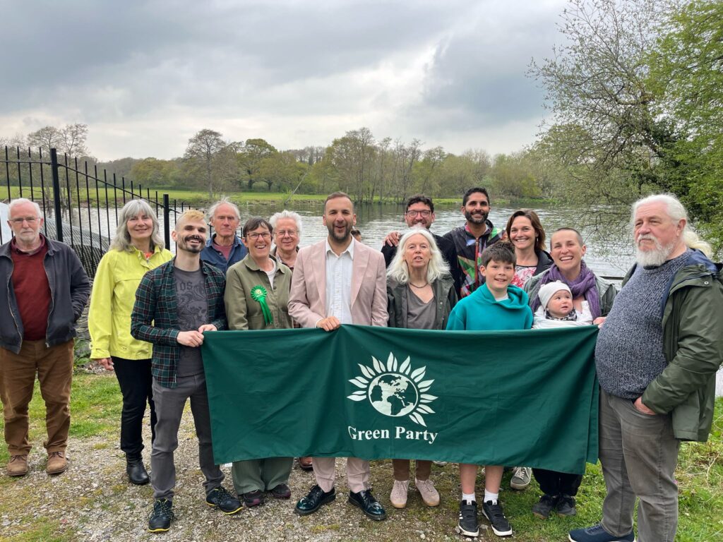 Zack Polanski stands with Green Party activists on the banks of the River Dart. They are holding a long Green Party banner. There are trees on the far bank and a cloudy sky.